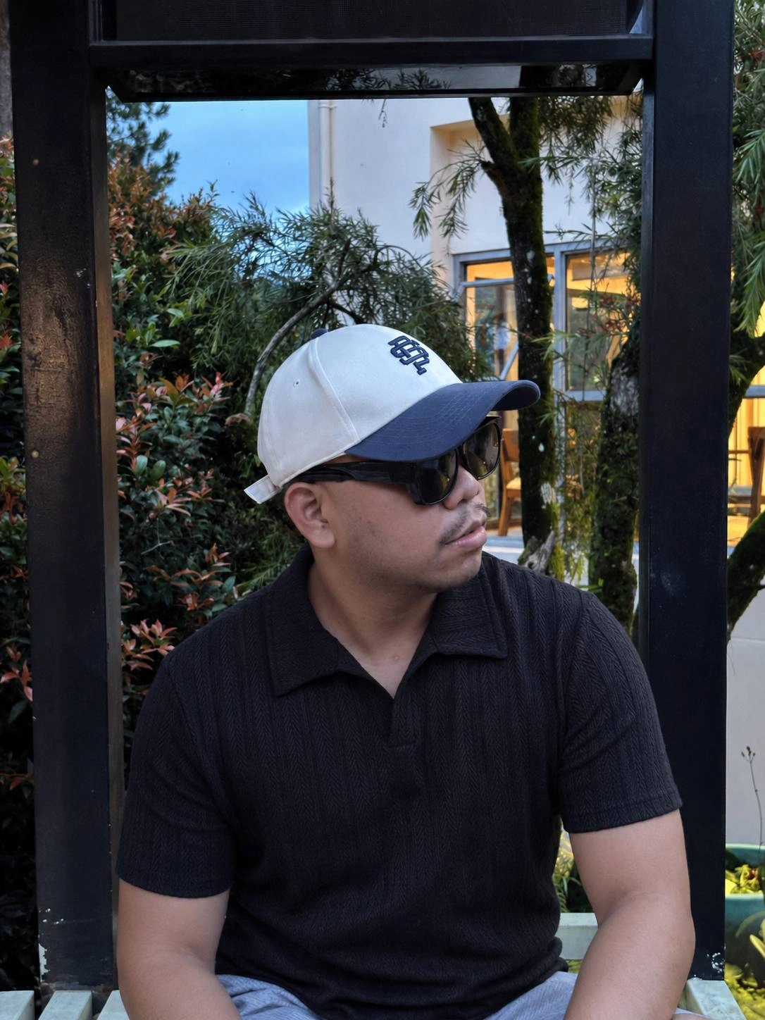 Den Jansen Flores in a black polo shirt, cap and sunglasses, seated inside a dark architectural frame.